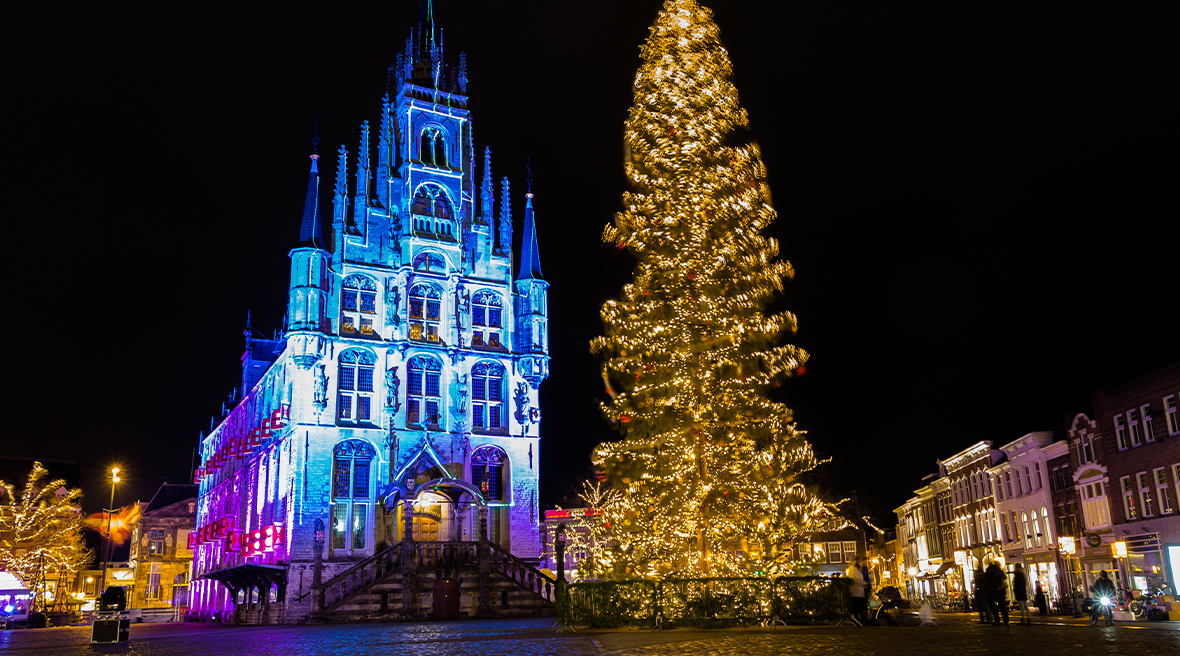 Huge brightly lit Christmas tree at night with Gouda town hall behind it, illuminated in light blue